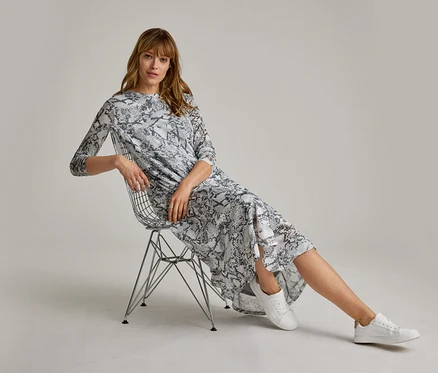 Femme en robe midi imprimé serpent gris et blanc assise sur une chaise en fil de fer, portant des baskets en cuir blanches.