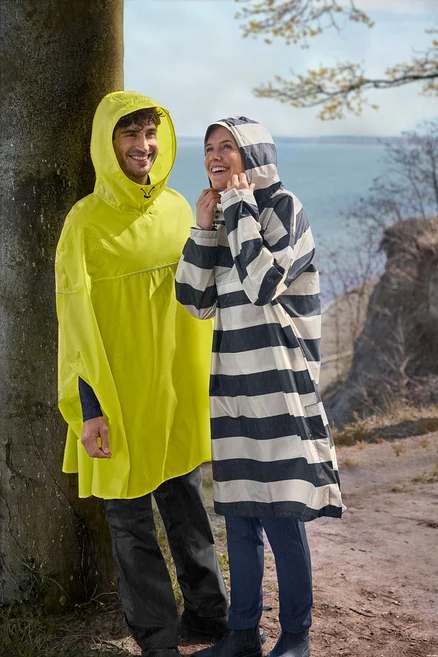 Homme en poncho de pluie jaune et femme en poncho de pluie rayé noir et blanc souriant dehors sous une légère pluie près d'un arbre et de l'eau.