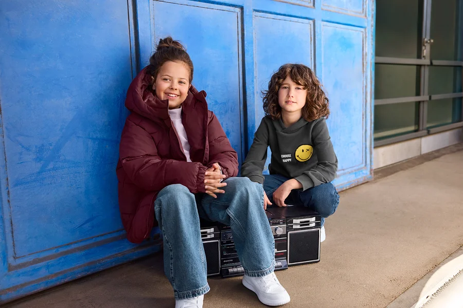 Deux enfants sont assis devant un mur bleu. Une fille en doudoune bordeaux sourit à gauche, tandis qu'un garçon en sweat à capuche gris avec un smiley est assis sur un boombox à droite.