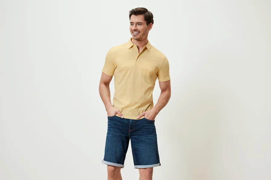 Un homme pose en polo en jersey jaune chiné et short en jean.