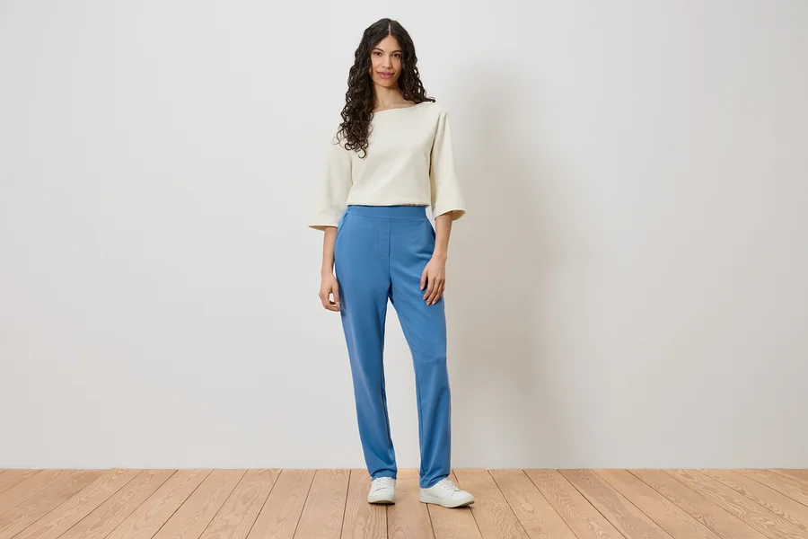 Une femme se tient devant un mur et porte un haut crème, un pantalon bleu et des chaussures blanches.