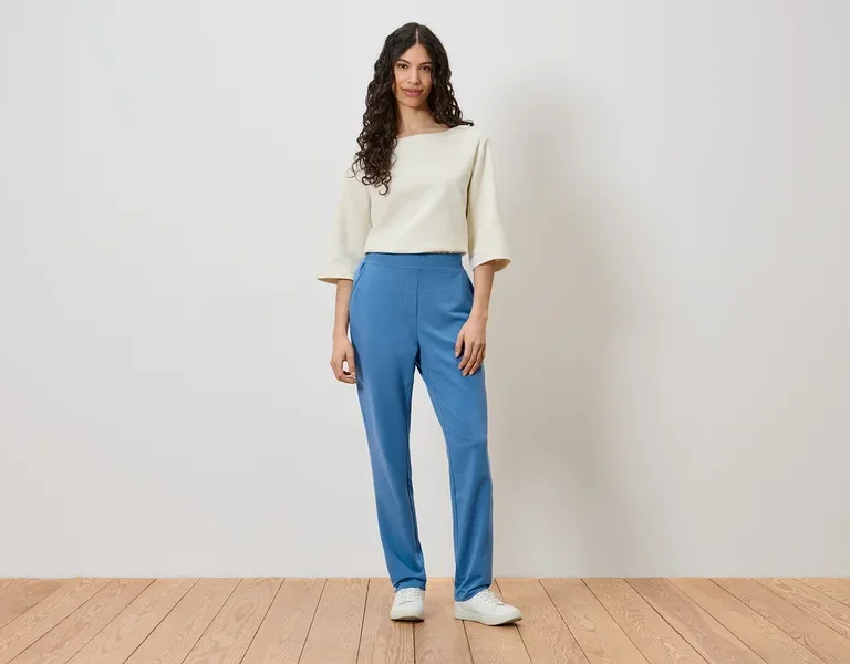 Une femme se tient devant un mur et porte un haut crème, un pantalon bleu et des chaussures blanches.