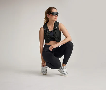 Femme en tenue de sport noire, gilet et lunettes de soleil accroupie sur fond clair.