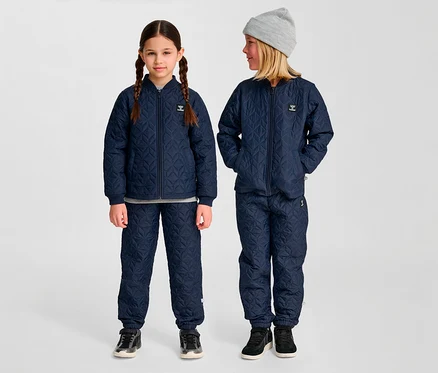 Deux enfants portent des ensembles hummel® « HMLSOBI Thermo-Set » bleus matelassés, composés d'une veste et d'un pantalon. Un enfant porte un bonnet gris.