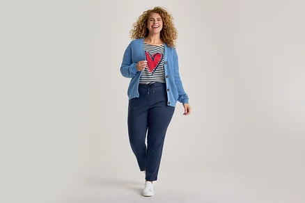 Une femme souriante aux cheveux bouclés marche vers l'avant, vêtue d'un cardigan bleu en maille, d'un t-shirt rayé avec un cœur rouge, d'un pantalon de jogging bleu marine et de baskets blanches.
