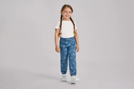 Une jeune fille aux deux tresses sourit en se tenant debout, vêtue d'un t-shirt blanc, d'un pantalon à enfiler pour enfant – Fit « Ella » bleu à motifs cœurs et de baskets blanches.
