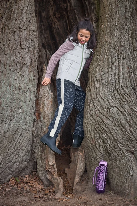 Une fille grimpe dans un tronc d'arbre creux, vêtue d'un gilet clair, d'un pantalon à motifs et de bottines Chelsea de pluie pour enfant bleu foncé. Une gourde violette est posée au sol.