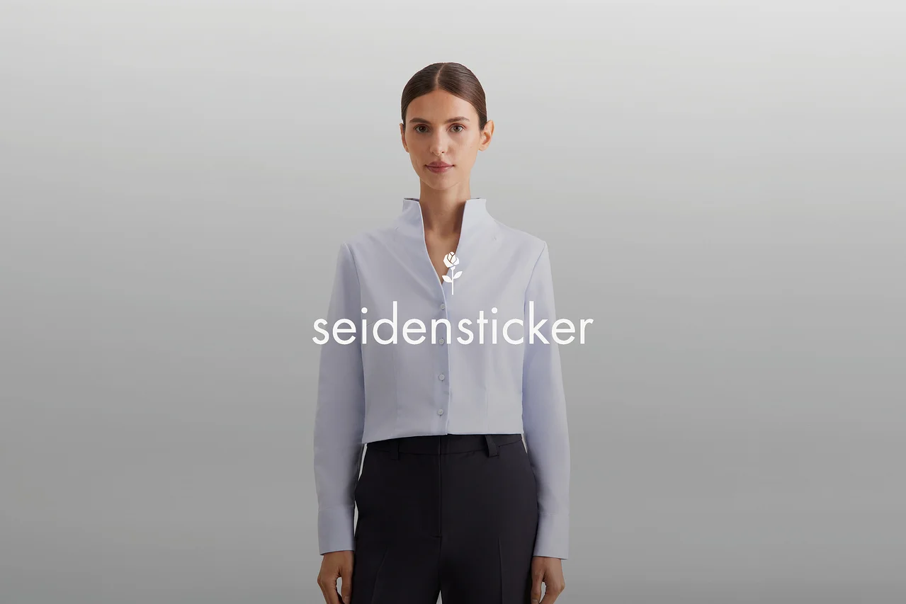 Une femme aux cheveux attachés porte une chemise courte bleu clair à col montant et un pantalon foncé. Un logo blanc "seidensticker" avec une rose est visible sur sa poitrine.