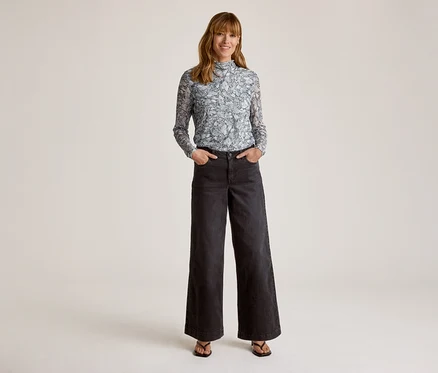 Femme souriante portant un haut à manches longues bleu clair à motifs et col montant, un pantalon large foncé et des sandales foncées, les mains dans les poches.