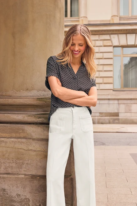 Une femme souriante aux longs cheveux blonds se tient les bras croisés, regardant vers le bas. Elle porte une chemise noire à motifs à manches courtes, un pantalon blanc à jambes larges et des Doubles créoles en or, appuyée contre un pilier en pierre à l'extérieur.