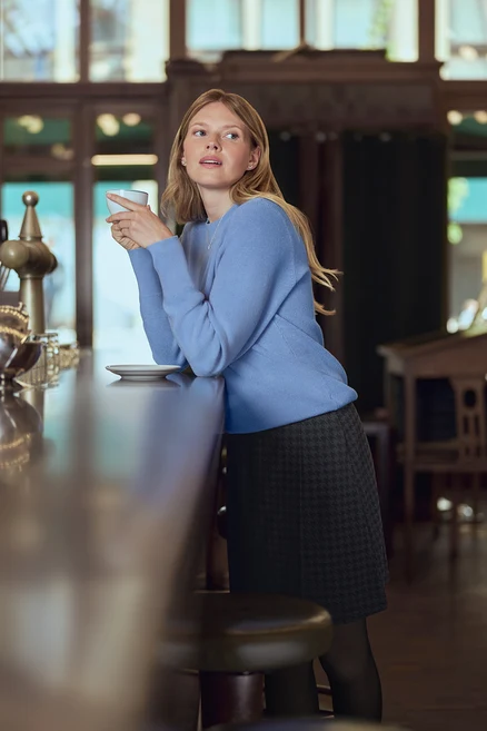 Une femme en pull bleu tient une tasse à un comptoir de bar. Elle porte des puces d’oreille avec arc en zircons en argent 925.