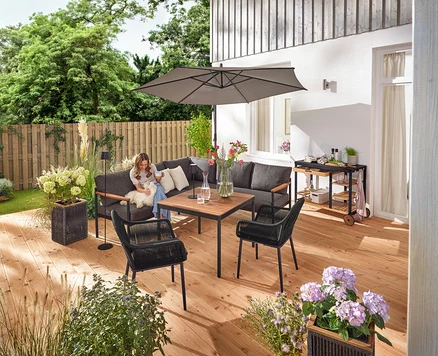 Sur une terrasse, une femme est assise avec un chien sur un coin salon-salle à manger »Elin«. On peut également voir un chariot de grill avec un plan de travail en polyciment, un pot de fleurs avec un tressage textile, un lampadaire d'extérieur LED sur batterie, une lampe de table d'extérieur LED sur batterie, deux coussins d'extérieur à toucher lin et une chaise de jardin avec un tressage croisé textile.