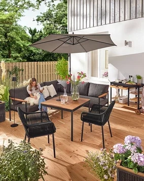 Sur une terrasse, une femme est assise avec un chien sur un coin salon-salle à manger »Elin«. On peut également voir un chariot de grill avec un plan de travail en polyciment, un pot de fleurs avec un tressage textile, un lampadaire d'extérieur LED sur batterie, une lampe de table d'extérieur LED sur batterie, deux coussins d'extérieur à toucher lin et une chaise de jardin avec un tressage croisé textile.