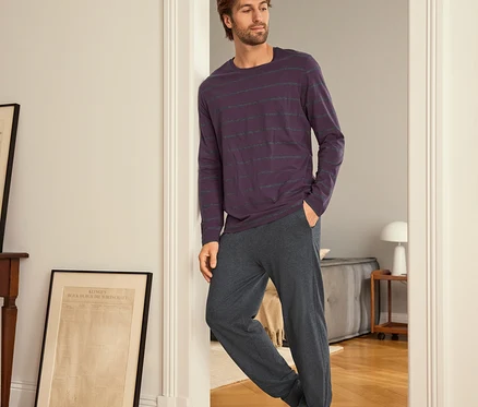 Un homme appuyé contre une porte, portant un t-shirt à manches longues rayé violet et un pantalon de survêtement gris.