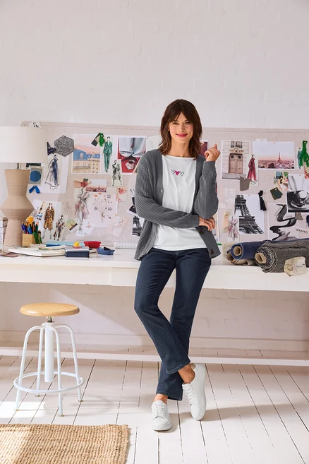 Femme souriante en cardigan gris, t-shirt blanc à cœurs, jean foncé et Baskets en cuir blanches, portant des Doubles créoles en or, devant un bureau avec un moodboard.
