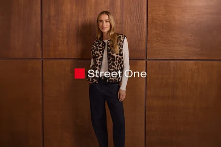 Une femme aux longs cheveux blonds se tient devant un mur en bois, vêtue d'un gilet imprimé léopard par-dessus un haut blanc à manches longues et d'un jean foncé. Elle a les mains dans les poches. Le logo "Street One" est visible.