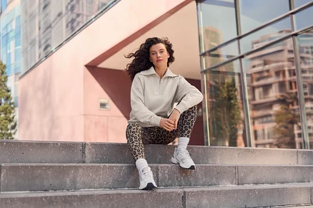 Femme aux cheveux bouclés assise sur des marches en béton, portant un haut beige à demi-zip, un legging imprimé léopard et des baskets blanches, avec des bâtiments modernes en arrière-plan.