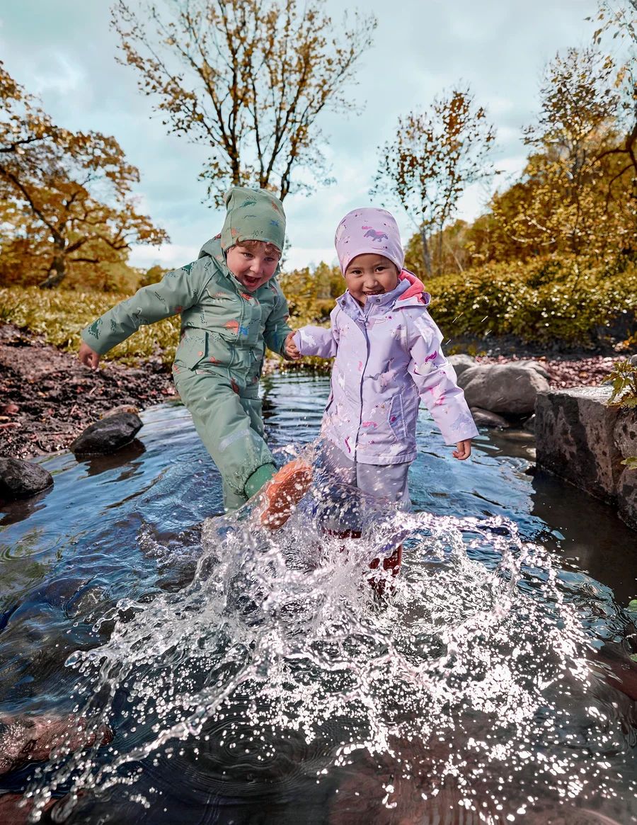 Deux jeunes enfants en tenues imperméables vertes et violettes éclaboussent l'eau dans un ruisseau peu profond, se tenant la main, avec des arbres d'automne en arrière-plan.