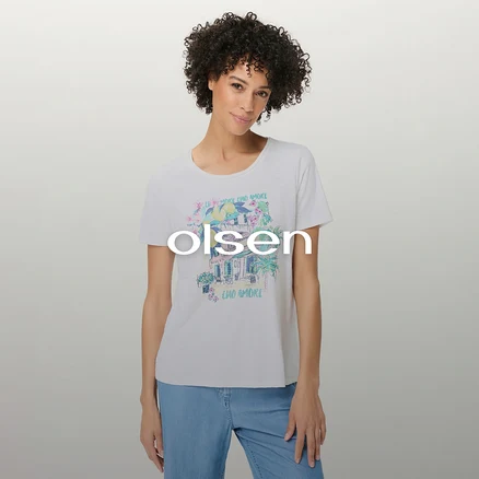 Femme aux cheveux bouclés portant un t-shirt blanc avec un imprimé graphique coloré d'un bâtiment, de fleurs, d'un palmier et du texte « CIAO AMORE », associé à un jean bleu clair.