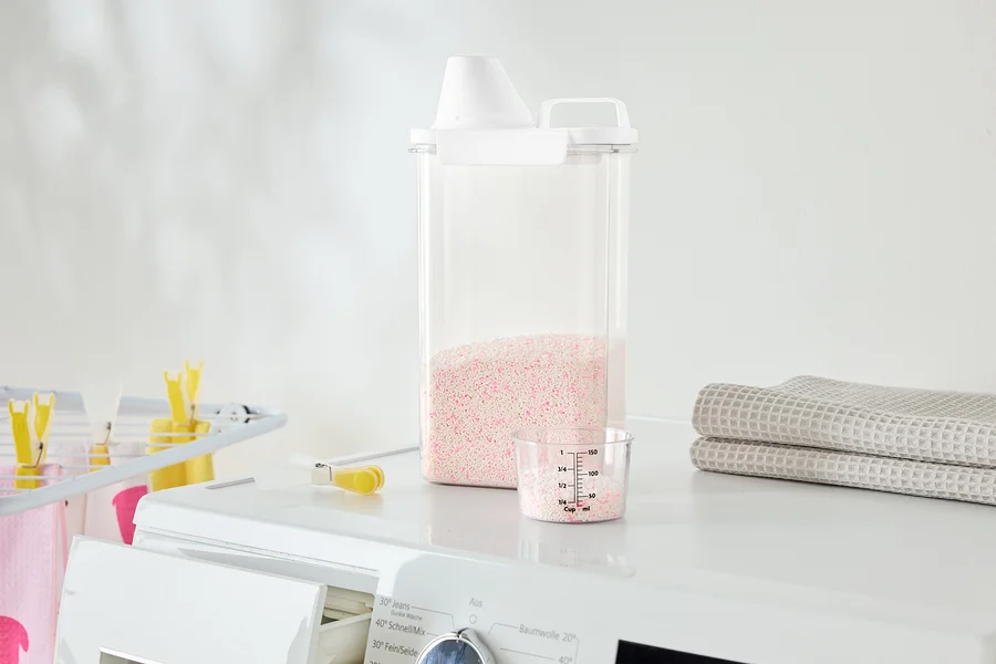 Sur un lave-linge se trouvent un récipient transparent avec de la lessive rose, un gobelet doseur et des serviettes pliées.