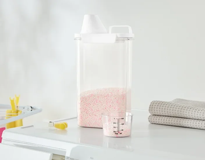 Sur un lave-linge se trouvent un récipient transparent avec de la lessive rose, un gobelet doseur et des serviettes pliées.