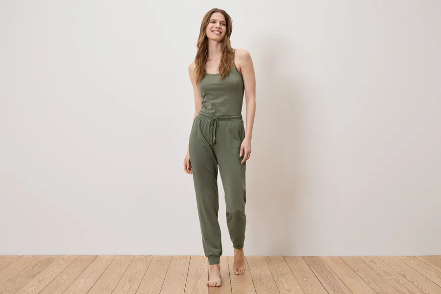Une femme se tient pieds nus sur un plancher en bois, portant un débardeur et un pantalon de jogging vert olive.