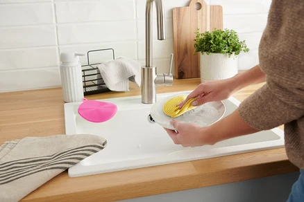 Une femme lave une assiette avec une éponge en silicone jaune dans un évier blanc. Une éponge rose est à côté.