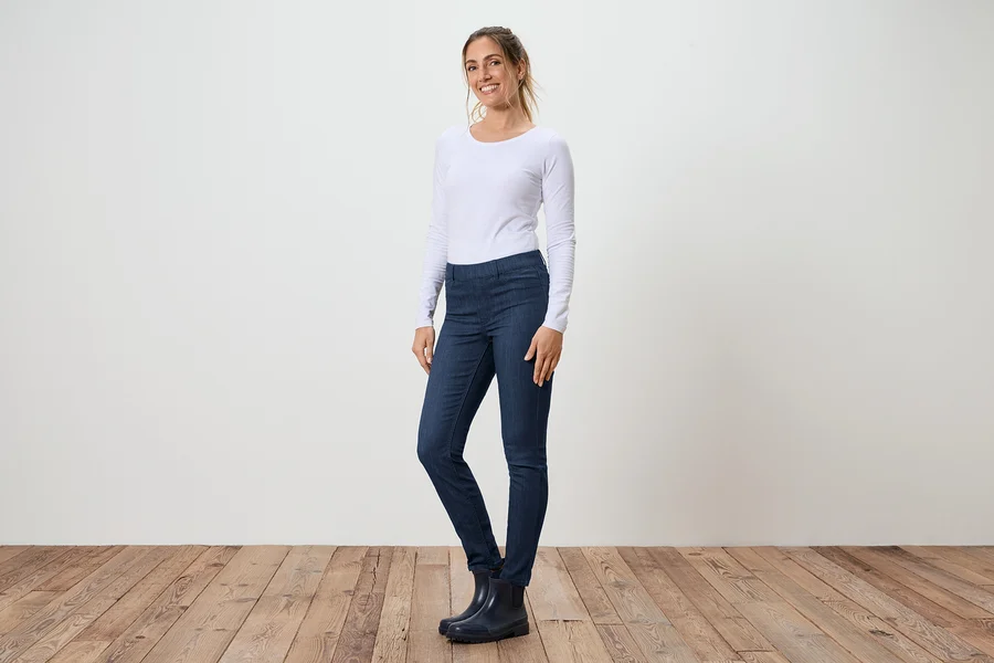 Une femme se tient de profil en jean, un T-shirt blanc à manches longues et des bottes en caoutchouc devant un mur blanc et un plancher en bois.