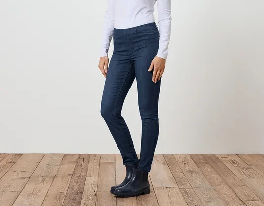 Une femme se tient de profil en jean, un T-shirt blanc à manches longues et des bottes en caoutchouc devant un mur blanc et un plancher en bois.