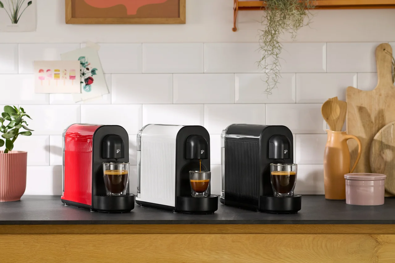 Trois machines à café rouges, blanches et noires sont placées côte à côte sur un comptoir de cuisine, chacune faisant couler du café dans une tasse placée en dessous.