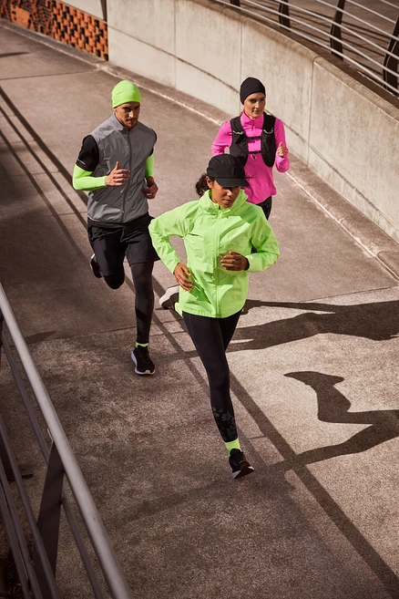 Trois coureurs, deux femmes et un homme, courent sur un chemin en béton. La femme à l'avant porte une casquette de sport noire, une veste de course neonlime, des leggings de sport « ActivePower » noirs et des chaussettes de course néon. L'homme au milieu porte un bonnet thermique neonlime, un gilet de course réfléchissant sur un t-shirt de sport noir et des manchettes de course à pied neonlime, un short de sport noir sur un collant de sport thermique et des chaussettes de course néon. La femme à l'arrière porte un bonnet thermique noir, un t-shirt de sport thermique rose avec demi-fermeture éclair et un gilet de course.