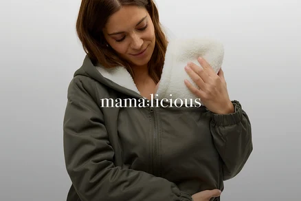 Une femme souriante aux cheveux bruns porte une veste verte doublée de polaire blanche et tient un bébé ou un insert de porte-bébé en fourrure blanche contre sa poitrine.