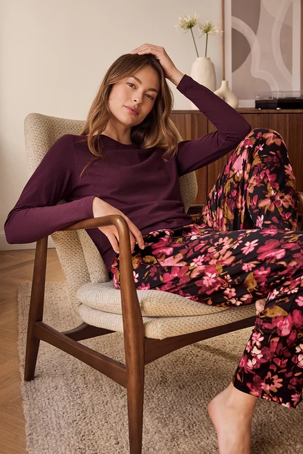 Femme assise confortablement dans un fauteuil clair, portant un haut à manches longues violet foncé et un pantalon noir à motif floral rose et rouge vif. Elle pose sa main sur sa tête et regarde devant elle. Un tapis shaggy est au sol, et une armoire en bois avec des décorations est en arrière-plan.