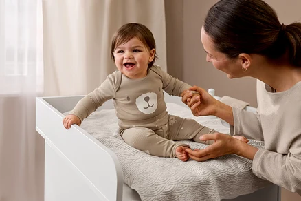 Bébé rieur en pull en maille fine beige chiné avec motif ours et pantalon assorti, assis sur la commode à langer « Leira », tenant la main d'un adulte.