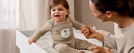 Bébé rieur en pull en maille fine beige chiné avec motif ours et pantalon assorti, assis sur la commode à langer « Leira », tenant la main d'un adulte.