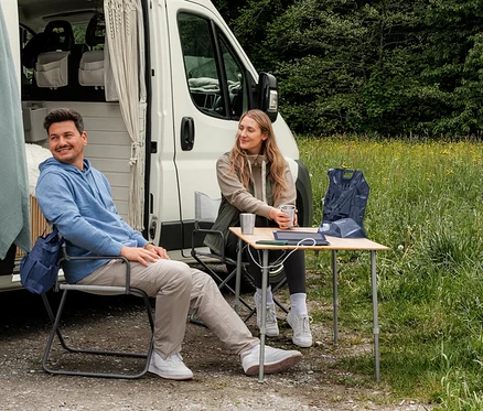 Un homme en sweat-shirt à capuche bleu clair et une femme en veste en sweat beige et gilet hybride kaki sont assis devant un camping-car. Un gilet pliable bleu foncé est posé sur une table.
