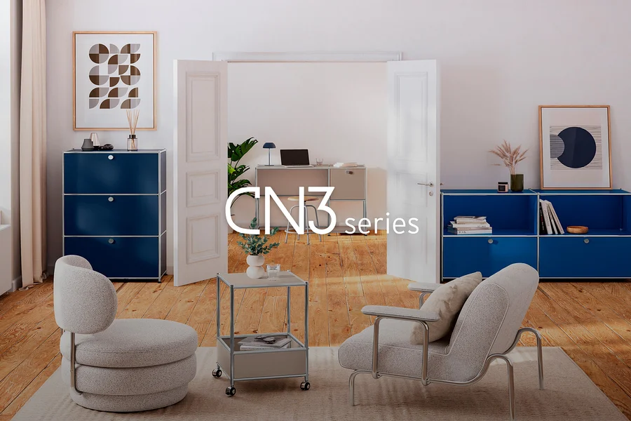 Un salon moderne avec deux fauteuils gris clair et une table d'appoint roulante sur un tapis. À gauche, une haute armoire modulaire bleu foncé à trois tiroirs, et à droite, une large armoire modulaire bleu foncé avec des étagères ouvertes et deux tiroirs, toutes deux décorées. À travers une porte ouverte, un bureau modulaire gris clair avec un ordinateur portable et une lampe est visible en arrière-plan. Texte superposé : CN3 series.