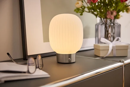 Une lampe de chevet avec un abat-jour blanc est posée sur une armoire à côté de lunettes, d'un livre, d'un vase de fleurs et d'une boîte cadeau.
