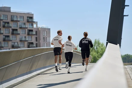 Trois personnes courent sur un pont: un homme en short de sport noir et T-shirt de sport blanc, une femme en leggings de sport « ActivePower » noirs et T-shirt de sport blanc, et une femme en sweatshirt noir et collant de sport « ActivePower » noir.