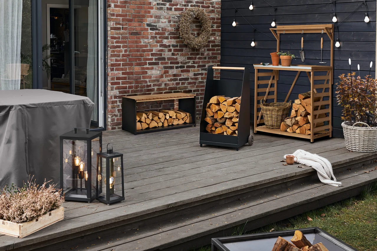 Une terrasse extérieure accueillante avec diverses solutions de rangement de bois de chauffage : un banc noir, un chariot et un établi de jardin en bois, tous remplis de bûches. Deux lanternes noires, une table recouverte de gris et une plante dans une boîte en bois sont à gauche. Une couverture blanche et une tasse reposent sur les marches, devant un mur de briques et de bois foncé.