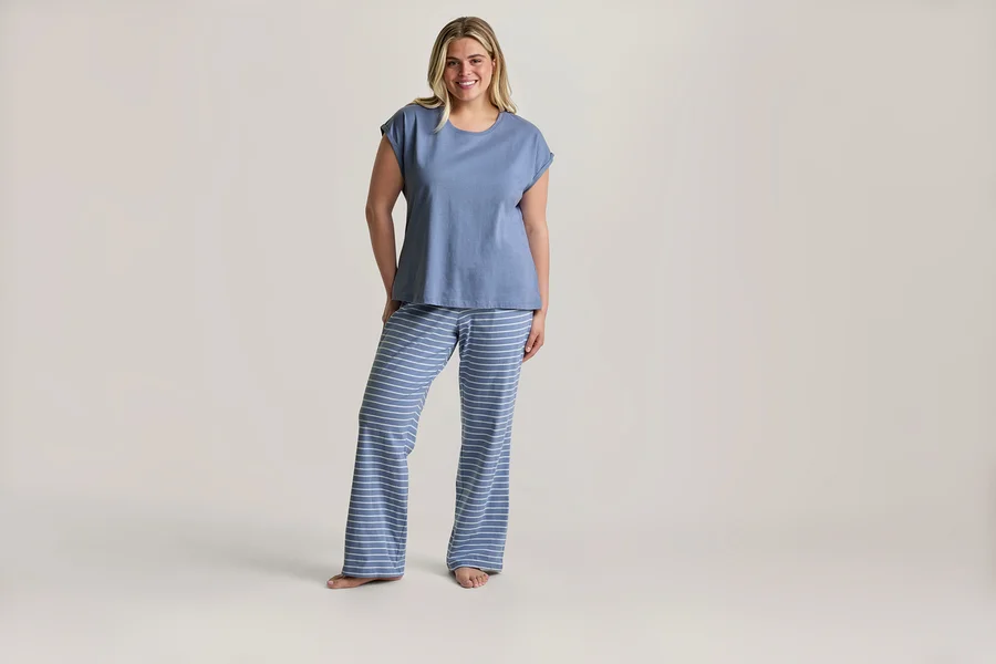Femme blonde souriante debout pieds nus, portant un ensemble de pyjama composé d'un t-shirt bleu et d'un pantalon rayé bleu et blanc.