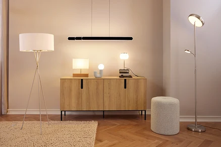 Une pièce moderne avec un buffet en bois surmonté de trois lampes et d'un objet décoratif, sous un luminaire suspendu linéaire noir. À gauche, un lampadaire trépied blanc repose sur un tapis, et à droite, un lampadaire halogène argenté avec liseuse se tient à côté d'un pouf gris.
