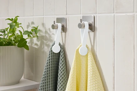 Deux serviettes, une grise et une jaune, sont accrochées à des crochets sur un mur carrelé. Une plante en pot est à gauche.