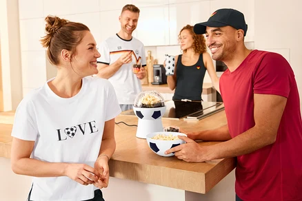 Quatre personnes dans une cuisine. Un homme tient un bol rempli de popcorn à côté d'une Machine à popcorn de football. Une femme porte un T-shirt à imprimé.