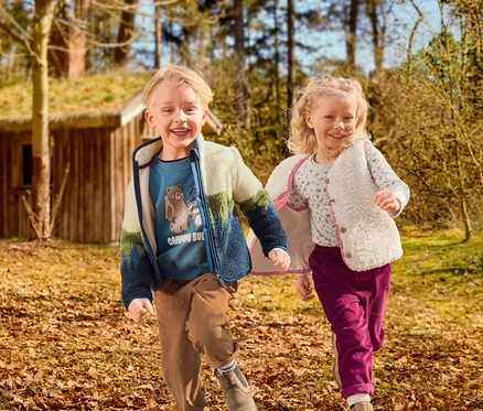 Deux enfants souriants, un garçon et une fille, courent dans un espace extérieur couvert de feuilles. Le garçon porte un t-shirt bleu à motif, une veste polaire colorée et un pantalon en velours côtelé marron. La fille porte un t-shirt à motifs, un gilet moelleux crème et un pantalon en velours côtelé magenta. Une maison en bois avec un toit en herbe est en arrière-plan.