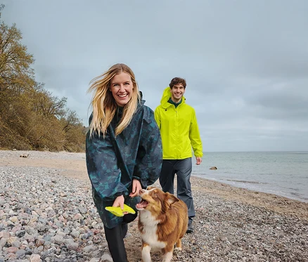 Une femme souriante en poncho de pluie foncé tient un frisbee jaune en marchant avec un chien marron et blanc sur une plage de galets. Un homme souriant en veste de pluie jaune marche derrière eux, avec la mer et un ciel nuageux en arrière-plan.