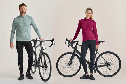 Un homme et une femme se tiennent à côté de vélos de route noirs. L'homme porte un maillot de cyclisme vert clair à manches longues et un pantalon de cyclisme noir. La femme porte un maillot de cyclisme rose foncé à manches longues et un pantalon de cyclisme noir.