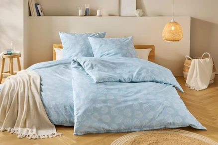 Une chambre avec une literie bleu clair à motifs de coquillages, un tabouret en bois avec une couverture et une lampe tressée.