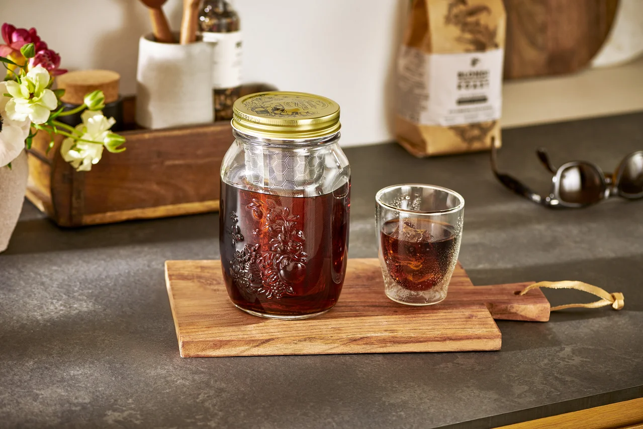 Café froid dans un bocal pour infuser le café à froid « BrewJar », à côté d'un verre sur une planche à découper en bois.