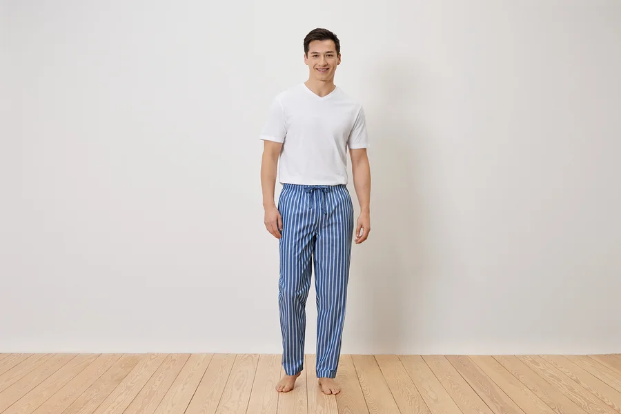 Un homme se tient pieds nus en pantalon de pyjama rayé bleu et blanc et en t-shirt blanc devant un mur blanc.